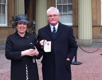 Des Evans and his wife stand in front of Buckingham Palace. He holds his OBE