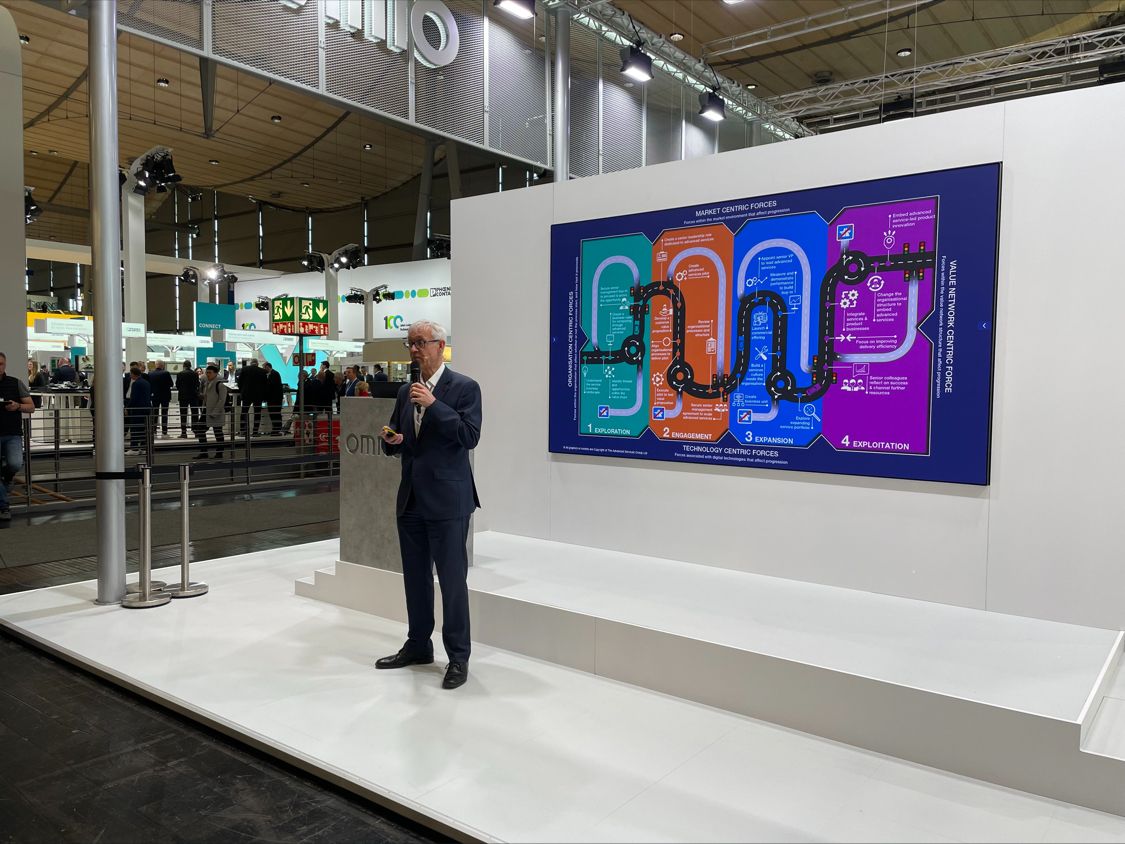 Market Centric Forces graphic sits on a white wall and in front of it, an Omron representative talks into a microphone