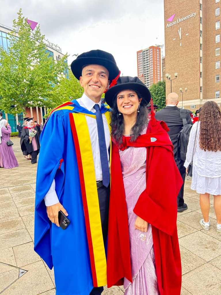 Two PHD graduates outside Aston University in graduating gowns and caps